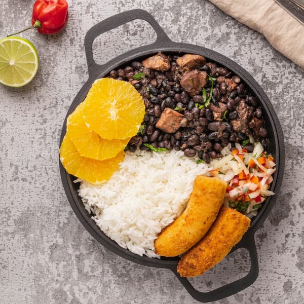 A black skillet holds Salsabor's black beans with meat, white rice, orange slices, and fried plantains.