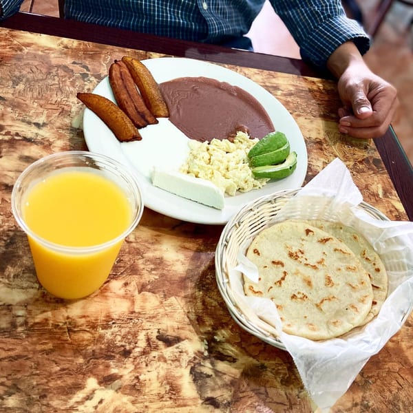 A Salvadoran breakfast with plantains, refried beans, scrambled eggs, cheese, avocado, tortillas, and orange juice.